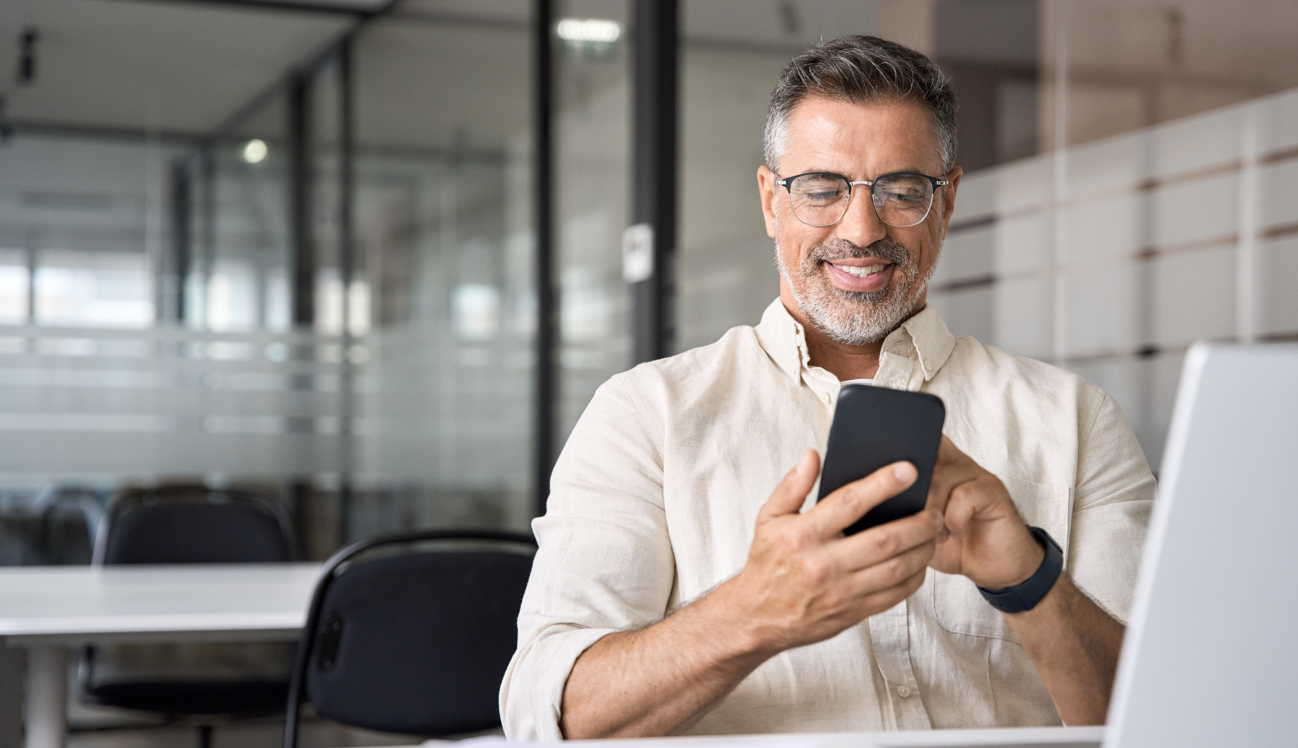 Middle aged Hispanic business manager ceo using cell phone mobile app, laptop. Smiling Latin or Indian mature man businessman holding smartphone sit in office working online on gadget with copy space.